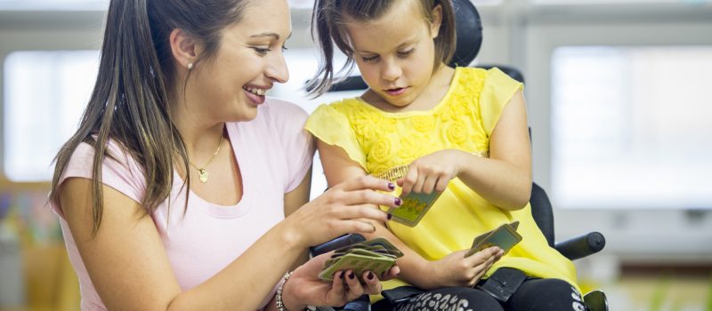 A Caucasian girl is sitting in her wheelchair inside a classroom. She is playing a card game with her educational assistant.